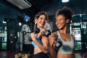 Two women smiling and holding dumbbells in a gym setting, showcasing strength training and fitness camaraderie, relevant to Vickery's Sweat Shop's focus on achieving fitness goals.